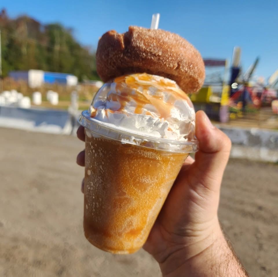 A hand holding a pumpkin cider slushie topped with whipped cream and an apple cider donut